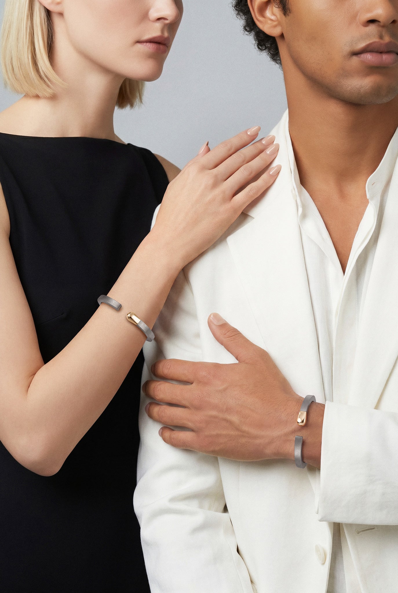 Models wearing Bracelets in 18k Rose Gold and grey titanium and a black dress for Her and a white suit for Him.